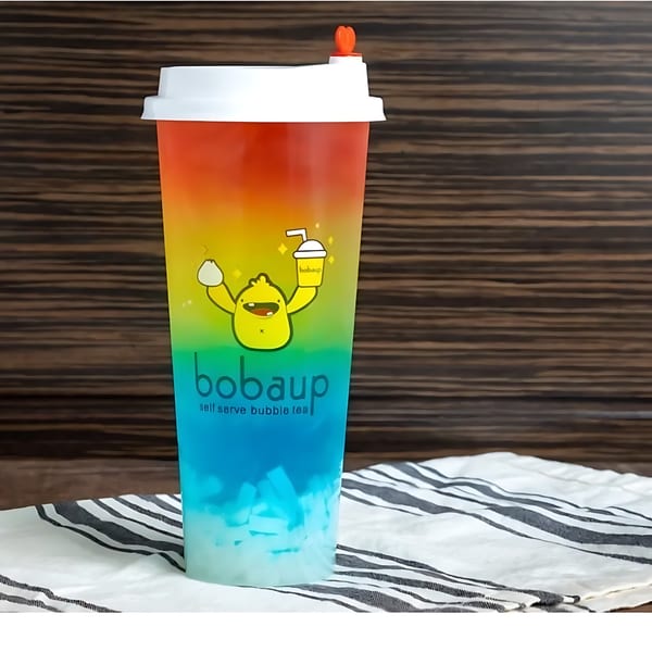 A Boba Up rainbow-colored boba drink with a cartoon character on a wooden table.