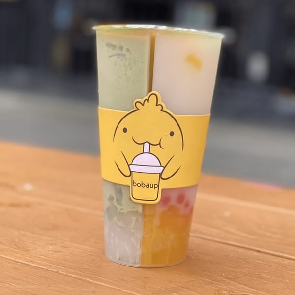 A Boba Up split cup with four different boba drinks and a yellow cartoon sleeve.