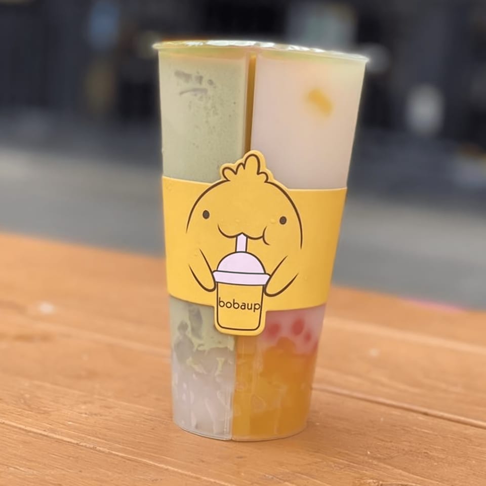 A Boba Up split cup with four different boba drinks and a yellow cartoon sleeve.