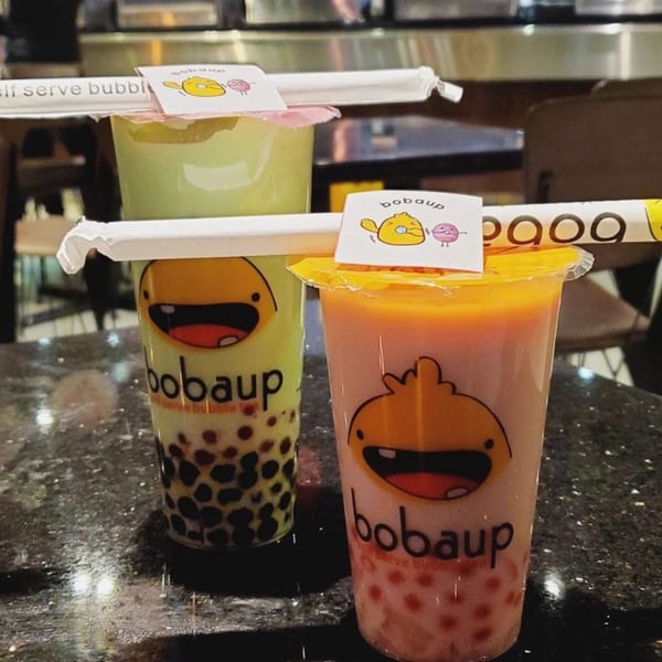 Two Boba Up drinks, one green with dark boba and one pink with red boba, on a dark counter.