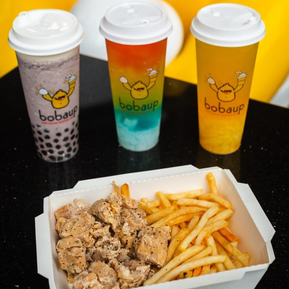Three Boba Up drinks and a box of fried chicken and french fries on a black surface.