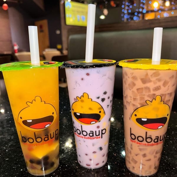 Three Boba Up bubble tea drinks: yellow, purple, and brown, with straws and cartoon logos.