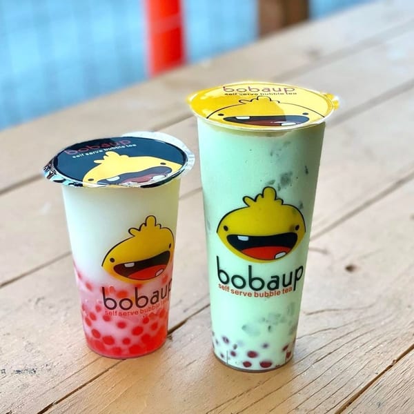Two Boba Up drinks, one pink with red boba and one green with dark boba, on a wooden table.