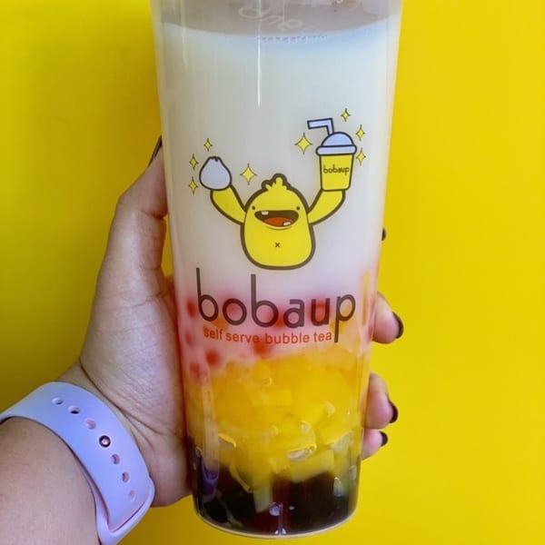 Hand holding a Boba Up bubble tea with layers of white milk, red, yellow, and dark brown toppings.