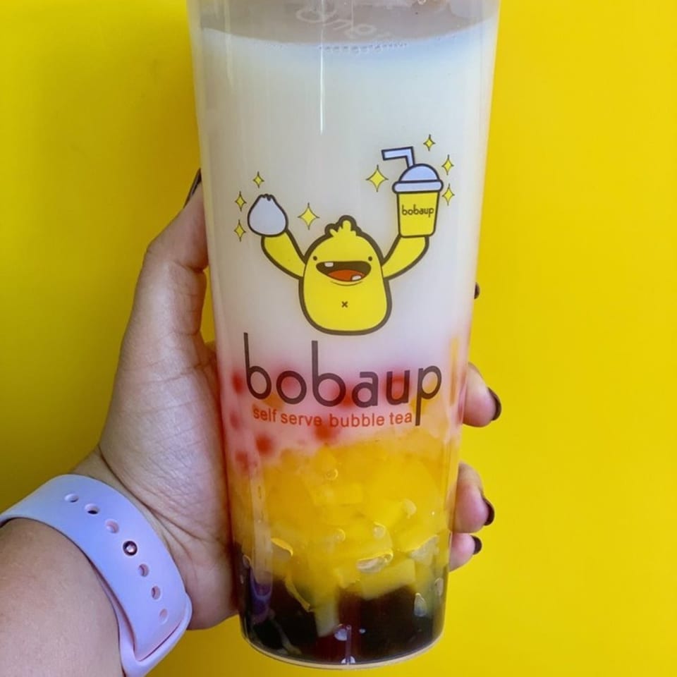 Hand holding a Boba Up bubble tea with layers of white milk, red, yellow, and dark brown toppings.