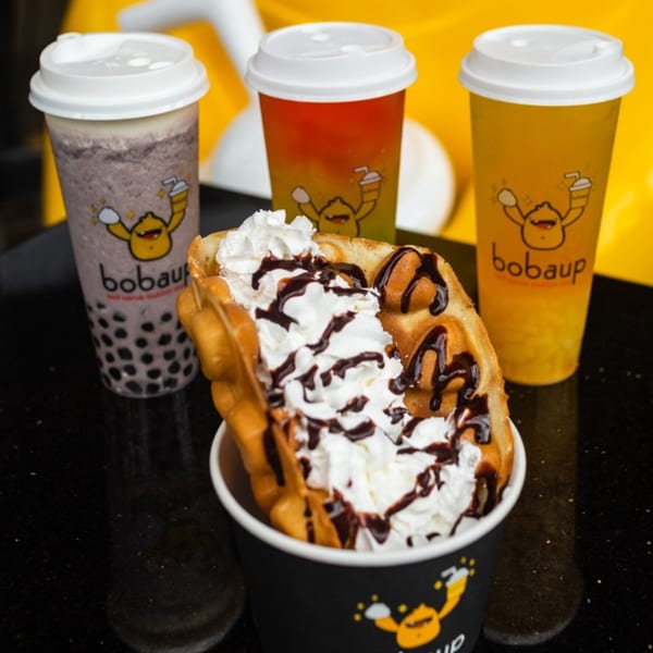 Boba Up drinks and a bubble waffle with whipped cream and chocolate drizzle on a black surface.